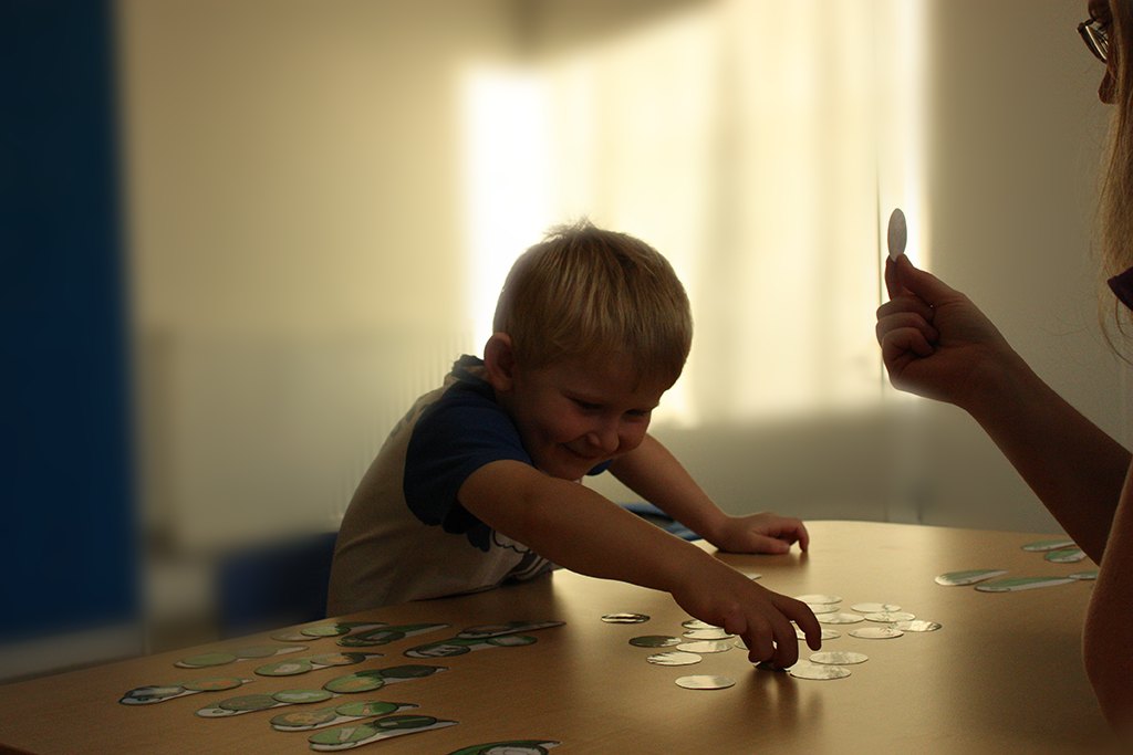 A young boy reaching across a table of picture cards, smiling, while Sarah holds another card up for him